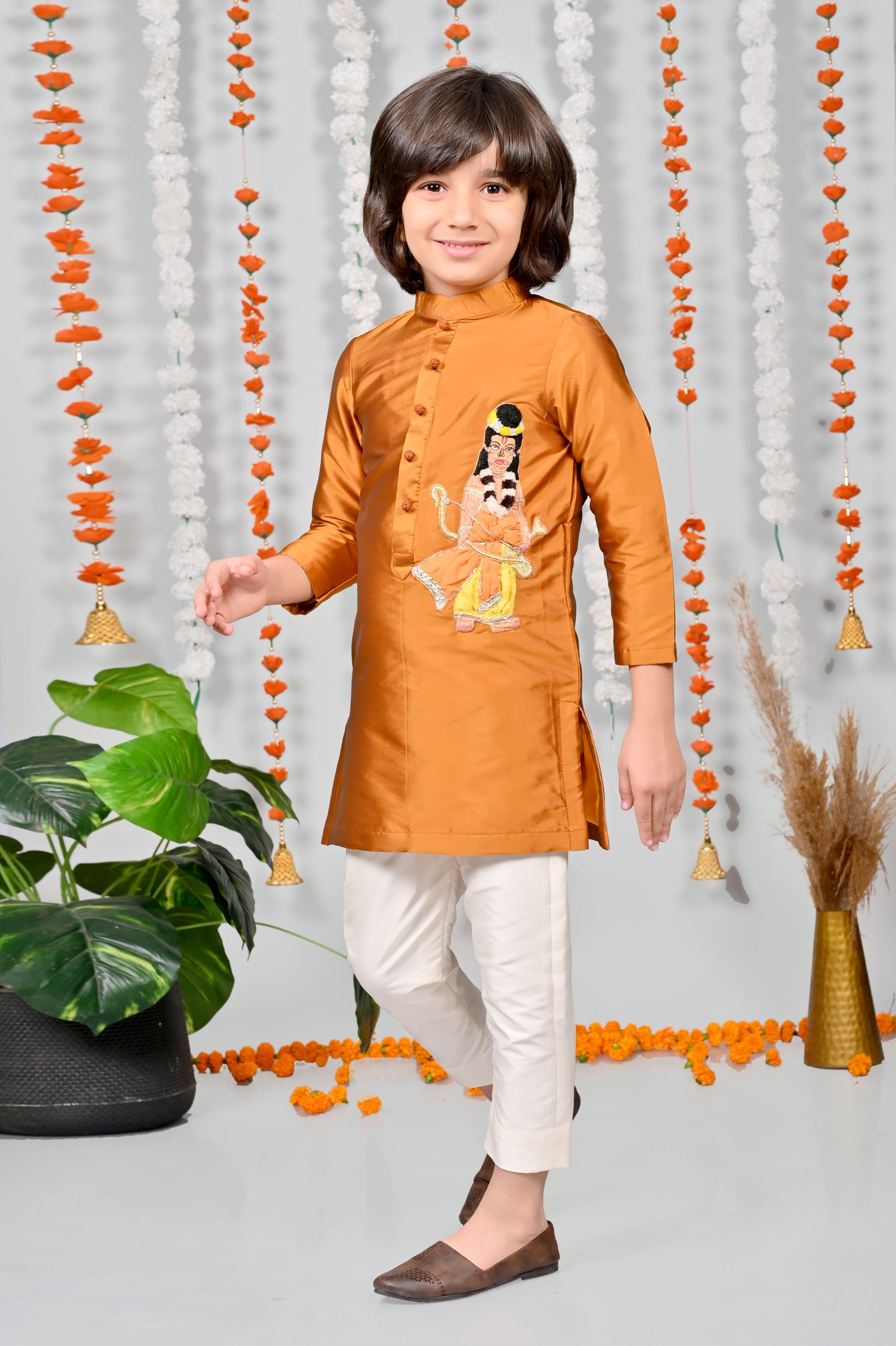 Child wearing an orange kurta with white pants standing against a decorative background.
